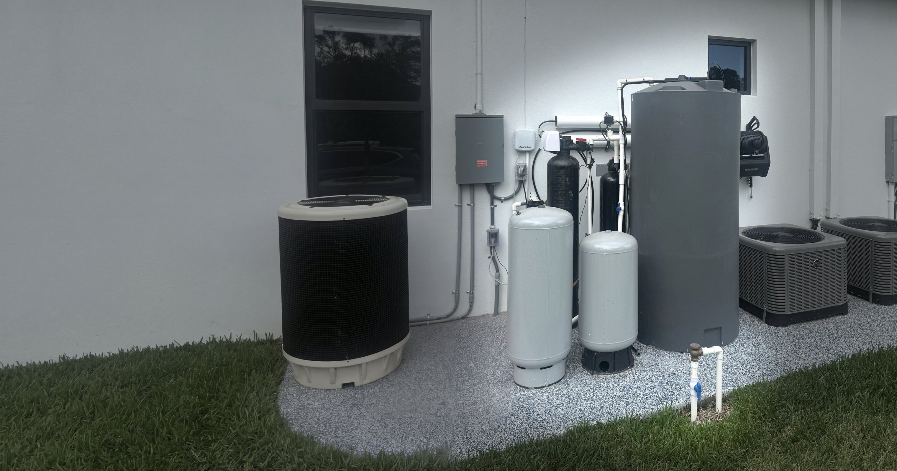 Whole-house water treatment system with a gray storage tank, filter tanks and 2 pressure tanks along the side of a white Florida home